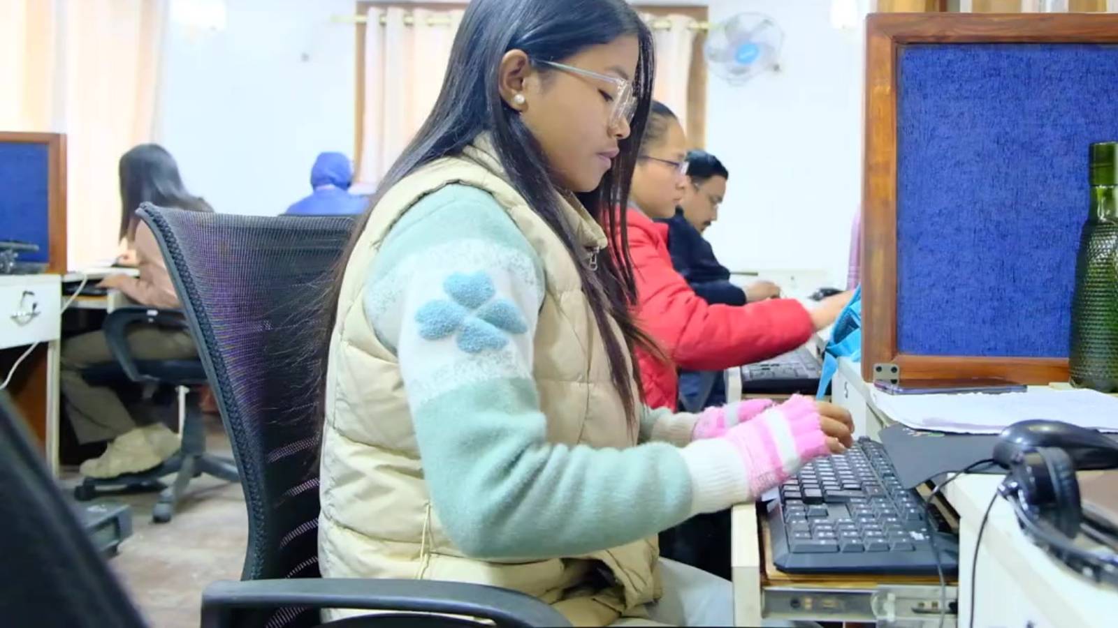 A woman in the foreground is typing at a computer. You can see other people behind her.