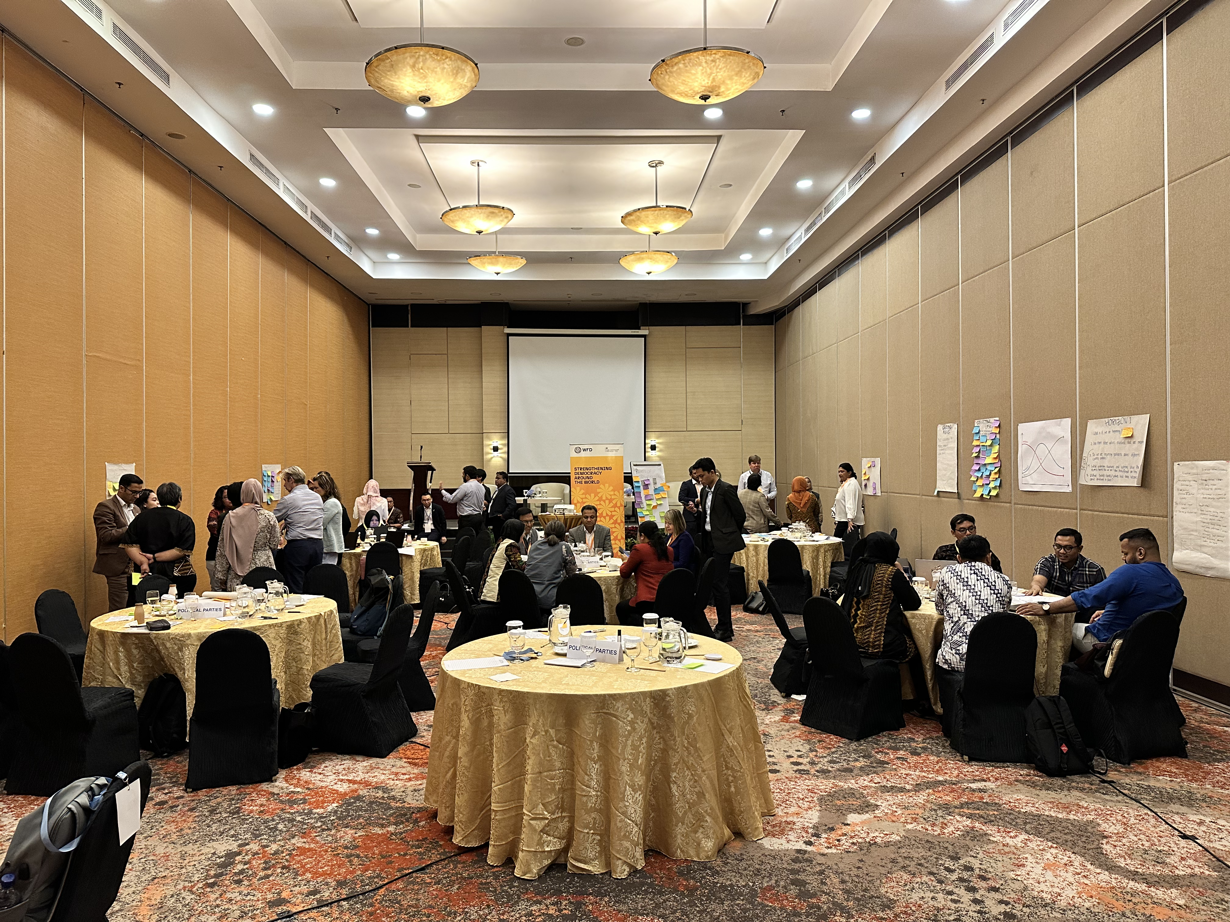 A photo of the Democracy Action Partnership event in action - a group of people are talking in groups, sat at tables or standing, concentration on brainstorming actions. In the centre is an orange banner which says strengthening democracy around the world.